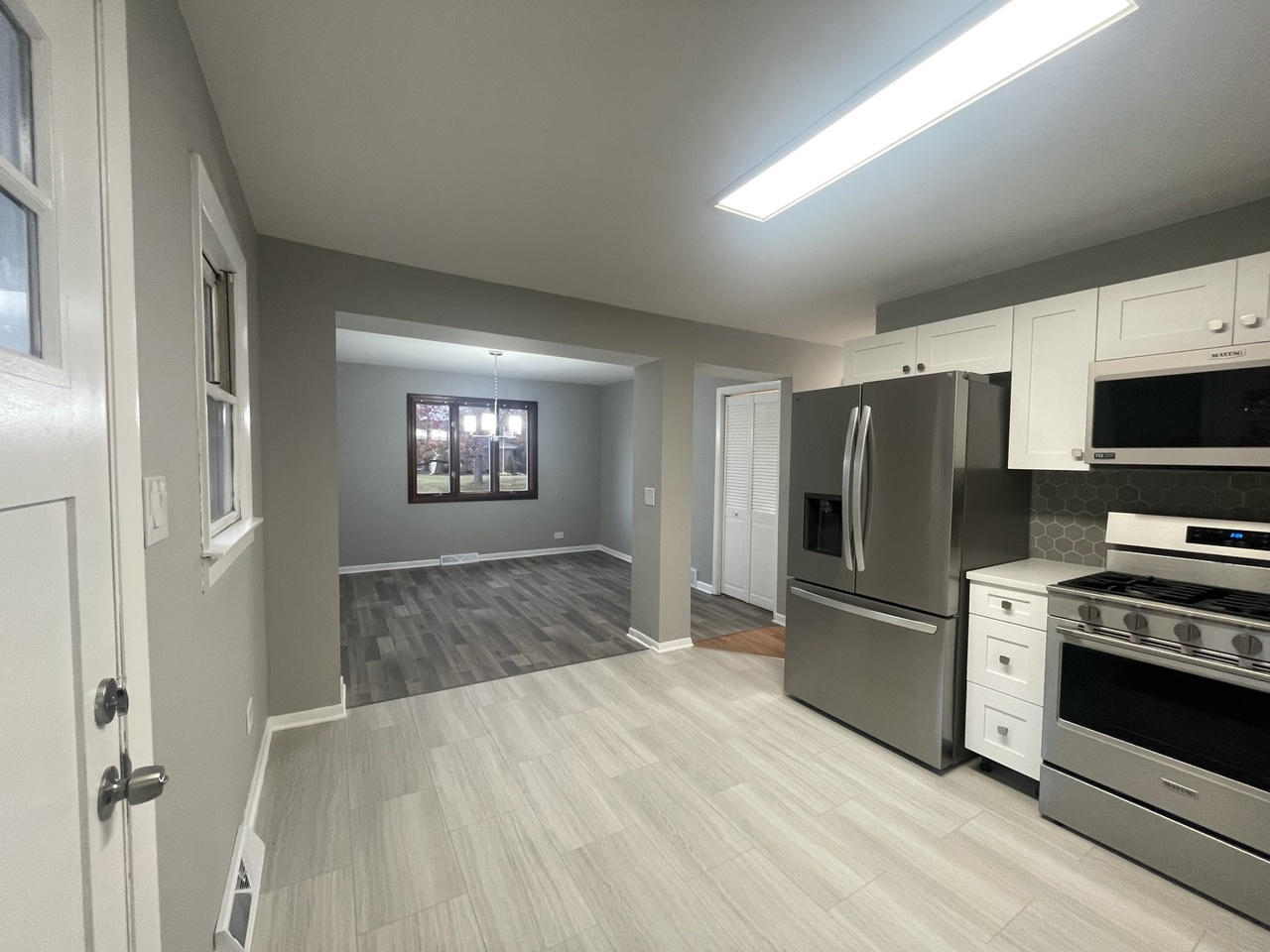 Modern kitchen with stainless steel refrigerator, stove, and microwave, white cabinets, gray backsplash, and beige tile flooring, leading to a living area with dark wood flooring and a window.