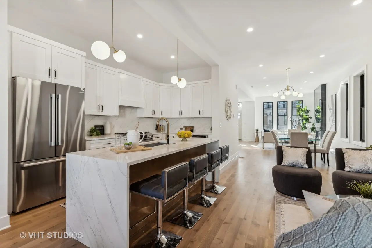 Modern open-concept kitchen and living room with white cabinets, marble countertops, stainless steel refrigerator, bar stools, dining table, armchairs, and large windows with plants.