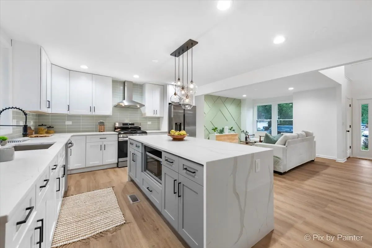 Modern open-concept kitchen with white cabinets, a kitchen island, and a view into a living room with large windows and a white sofa.