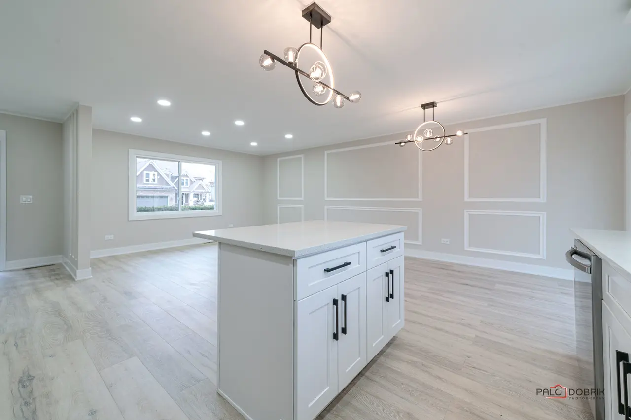 Modern kitchen with white island, light wood flooring, recessed ceiling lights, two contemporary chandeliers, and a window overlooking houses outside.