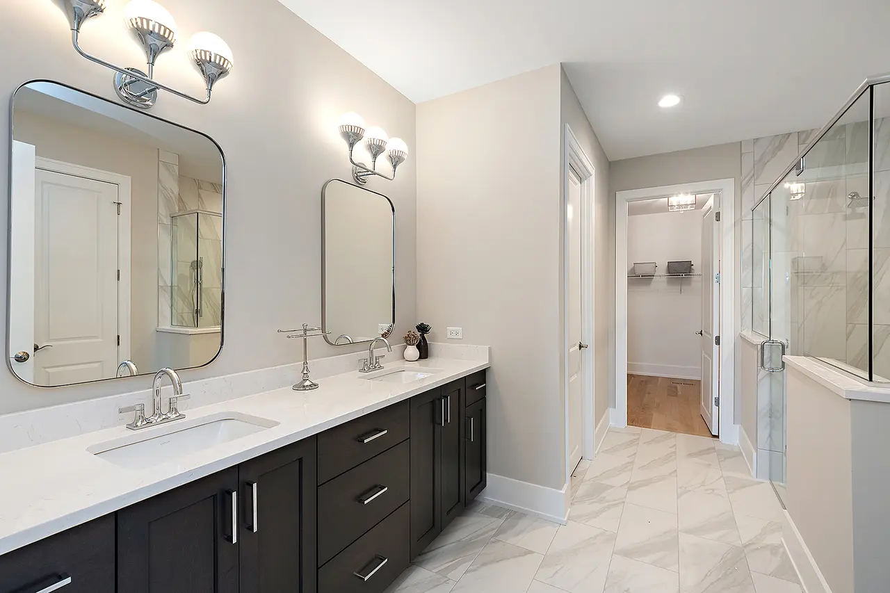 Modern bathroom with dual sinks, dark cabinetry, large mirrors, and a walk-in shower with glass enclosure and marble tiles.