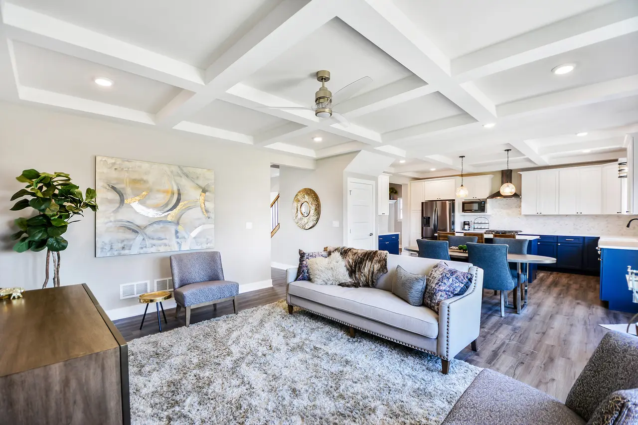 Open-concept living room and kitchen with modern decor, white walls, coffered ceiling, gray rug, gray sofa with throw pillows, blue chairs at dining table, stainless steel appliances, and wooden flooring.