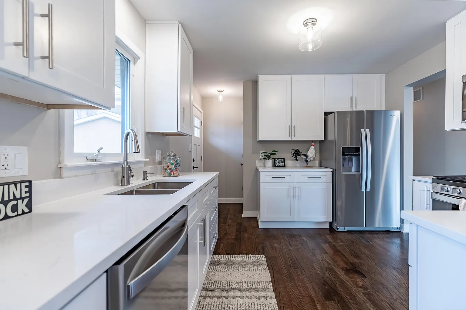 Modern kitchen with white cabinets, stainless steel appliances, and hardwood flooring. Features a double sink under a window, a refrigerator, and decorative items on the counter.