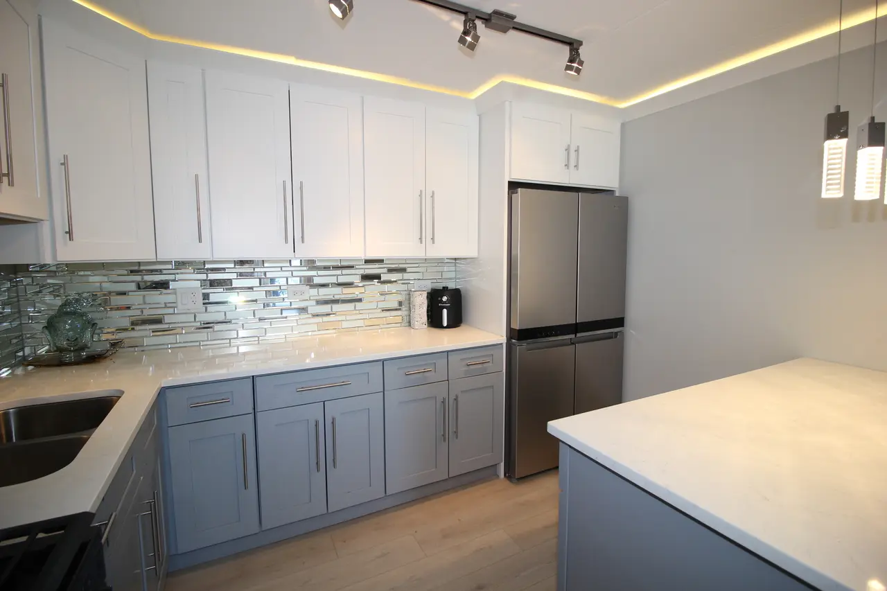 Modern kitchen with white upper cabinets, light blue lower cabinets, stainless steel refrigerator, tile backsplash, and a light-colored countertop. Ceiling features recessed lighting and track lights.
