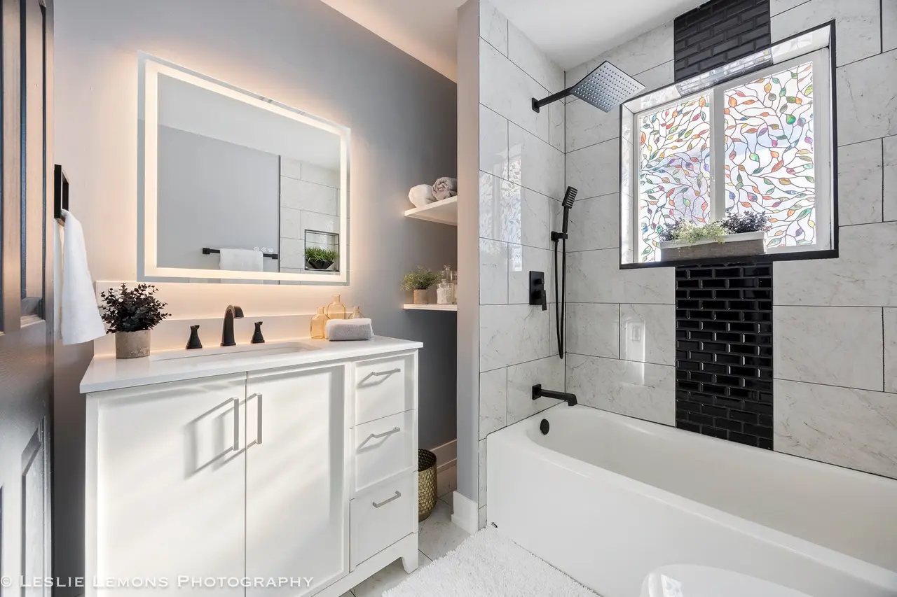 A modern bathroom with a vanity, mirror, and closet on the left, and a walk-in shower with a black rain shower head, a handheld shower, and a window with a colorful stained glass design on the right.