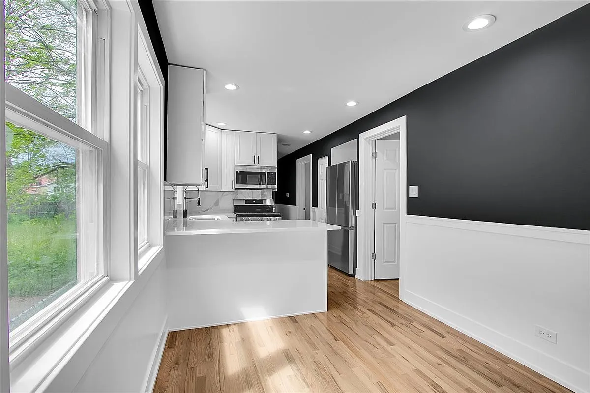 Modern kitchen with white cabinets, stainless steel appliances, and dark accent wall. Large window lets in natural light, hardwood floors throughout.