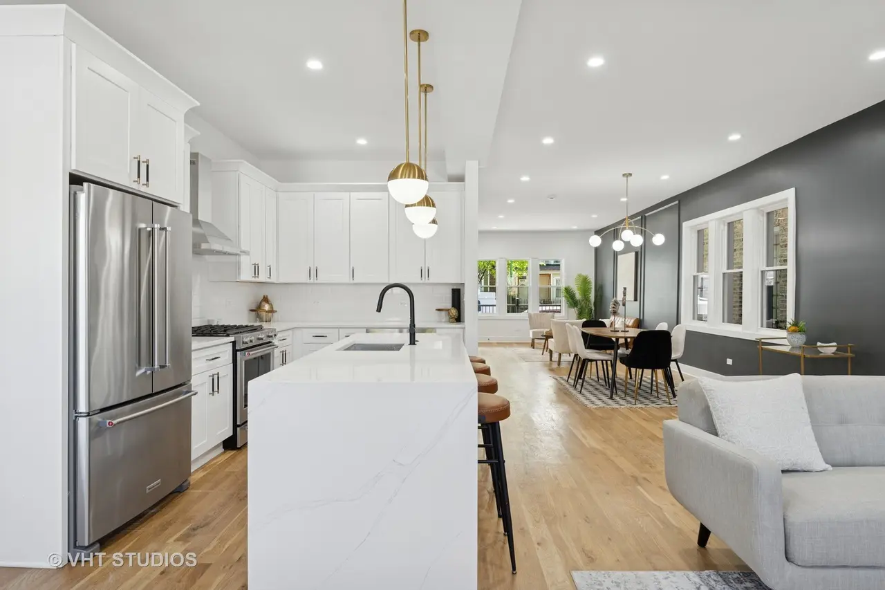 Open-concept modern kitchen and dining area with white cabinets, stainless steel refrigerator, and a seating island, adjacent to a living room with a light gray sofa, black and white chairs, and large windows.