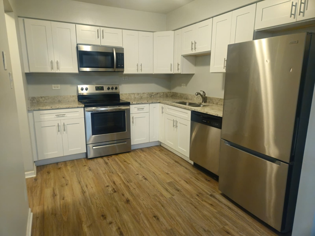 Kitchen with white cabinets, stainless steel refrigerator, stove, microwave, dishwasher, granite countertops, and wood flooring.