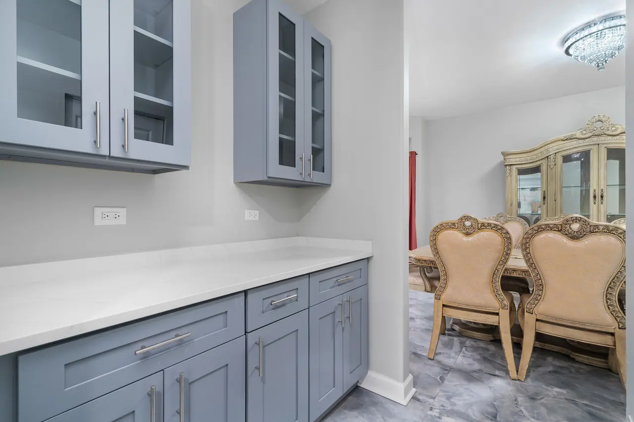 A corner of a modern kitchen with blue cabinets and white countertops, adjacent to a dining area with ornate chairs and a china cabinet in the background.