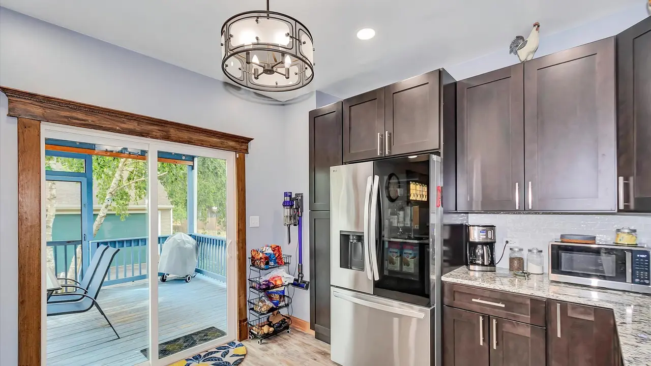 Kitchen with dark brown cabinets, stainless steel refrigerator, microwave, coffee maker, and countertop with jars, overlooking a deck outside through sliding glass doors.