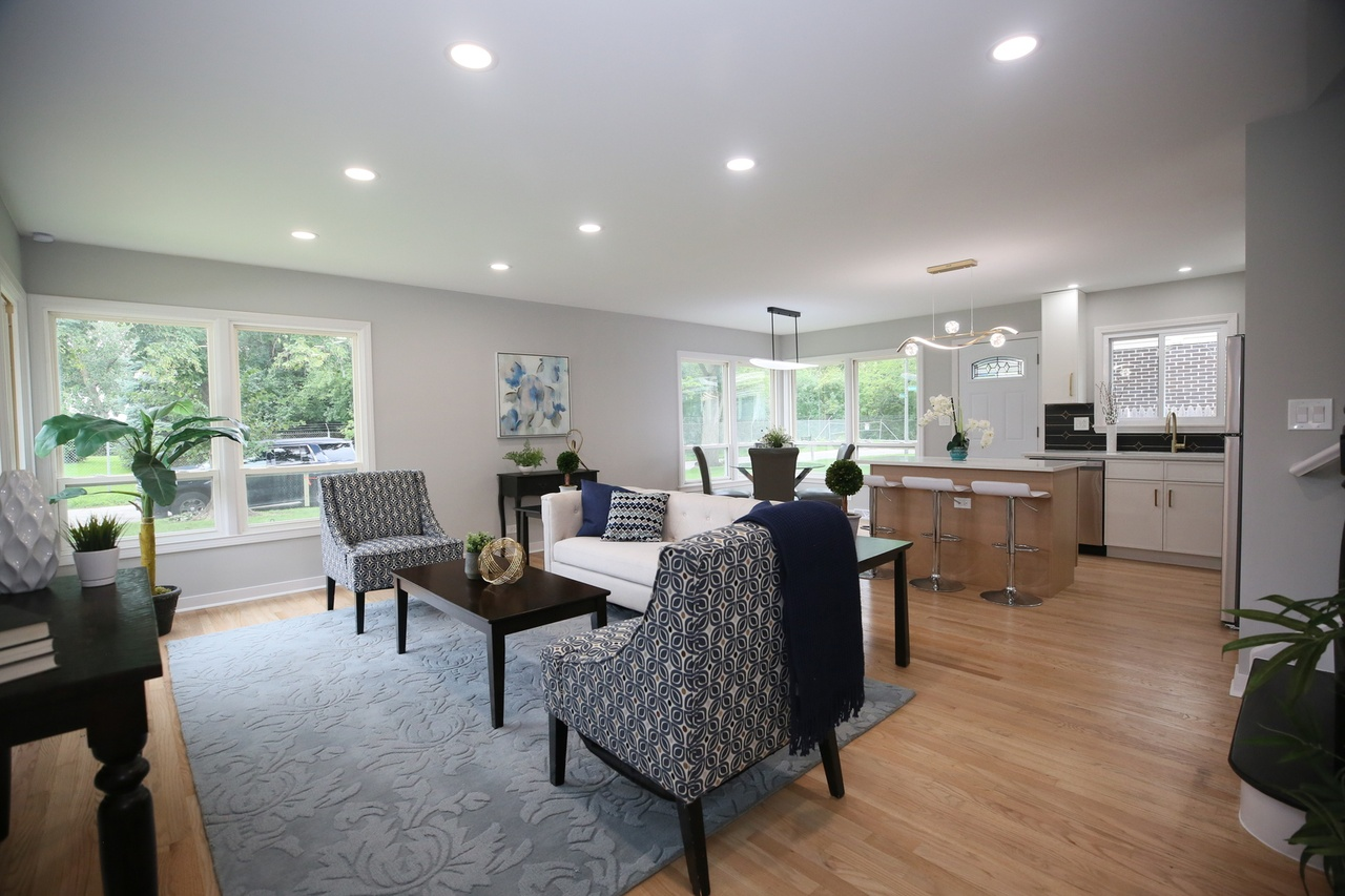 Open-concept living room and kitchen with hardwood floors, white walls, large windows, modern furniture, and pendant lighting.