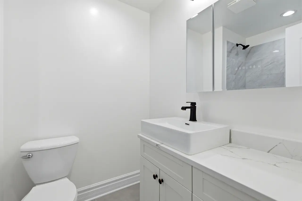 A clean, modern bathroom with a white toilet, white vanity with a vessel sink, black faucet, and a large mirror. In the reflection of the mirror, a shower with a black showerhead and marble tiles is visible.