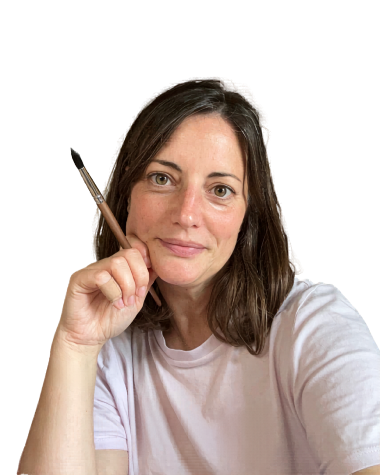 A woman with brown hair and fair skin, wearing a white t-shirt, holding a paintbrush, looking at the camera with a slight smile.