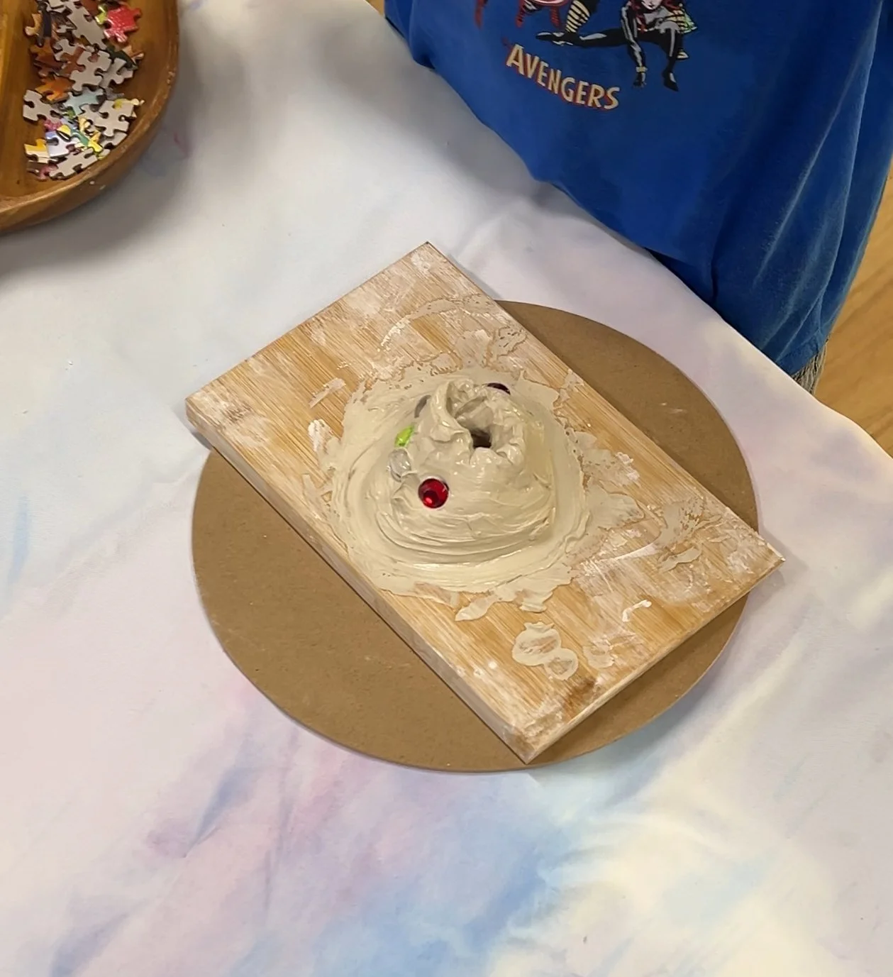 A wooden board with some white cream and small colorful candies or decorations on top, set on a round cardboard base, on a white tablecloth.