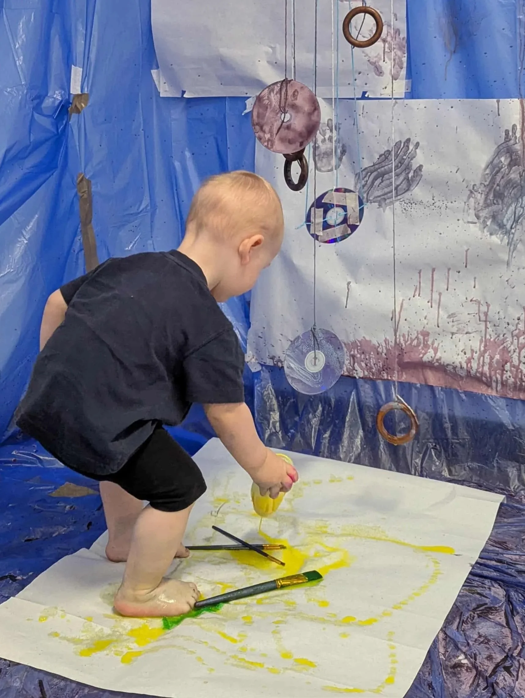 A young child with red hair is standing and playing with yellow paint on a large sheet of paper on the floor while holding a yellow container. The background has a blue plastic sheet with hanging metal and cardboard objects, creating an artistic or craft environment.