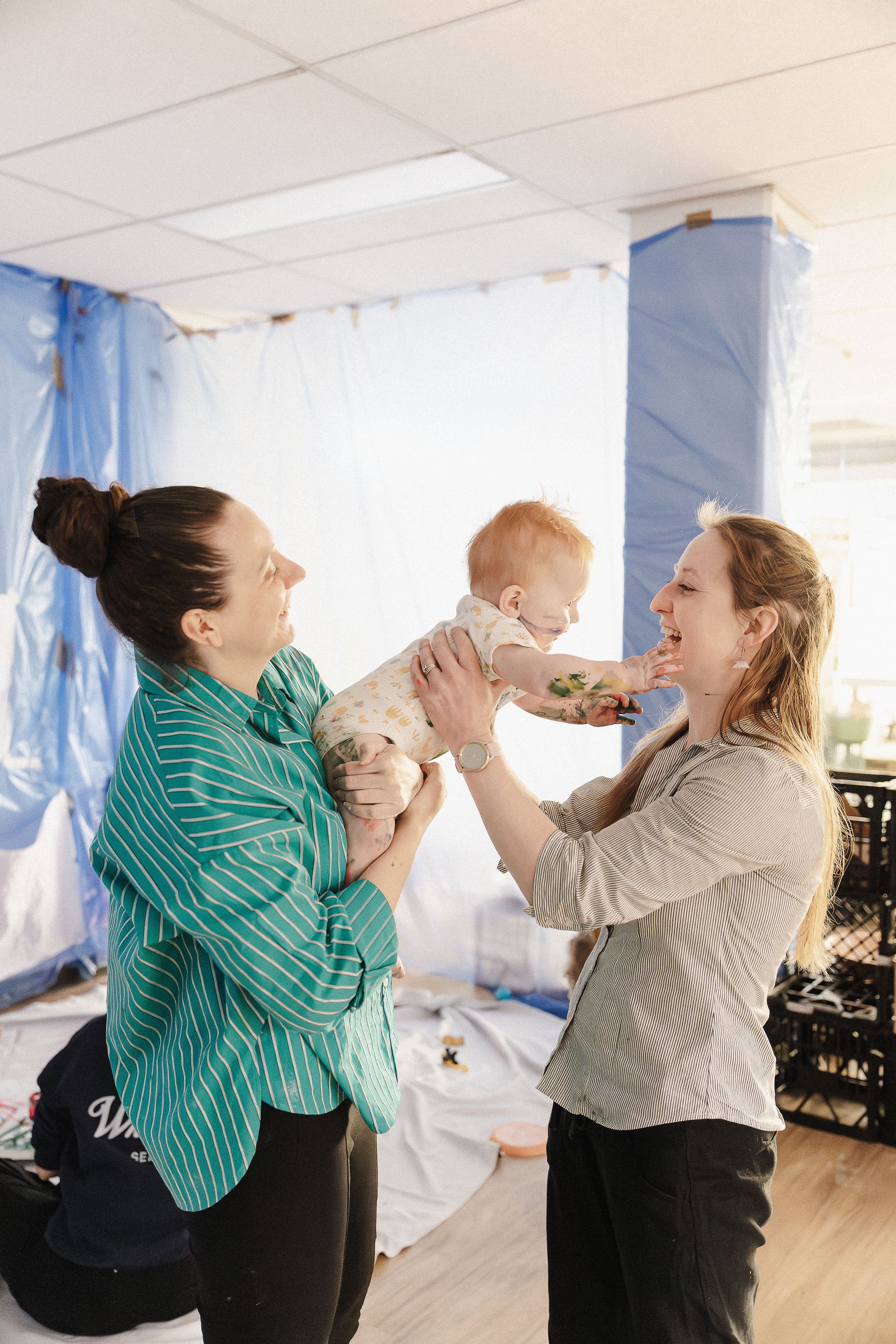 Two women playing with a young girl in a room with blue plastic sheets on the walls. The girl is reaching out to touch one woman's face while the other woman holds her. The scene appears joyful and playful.
