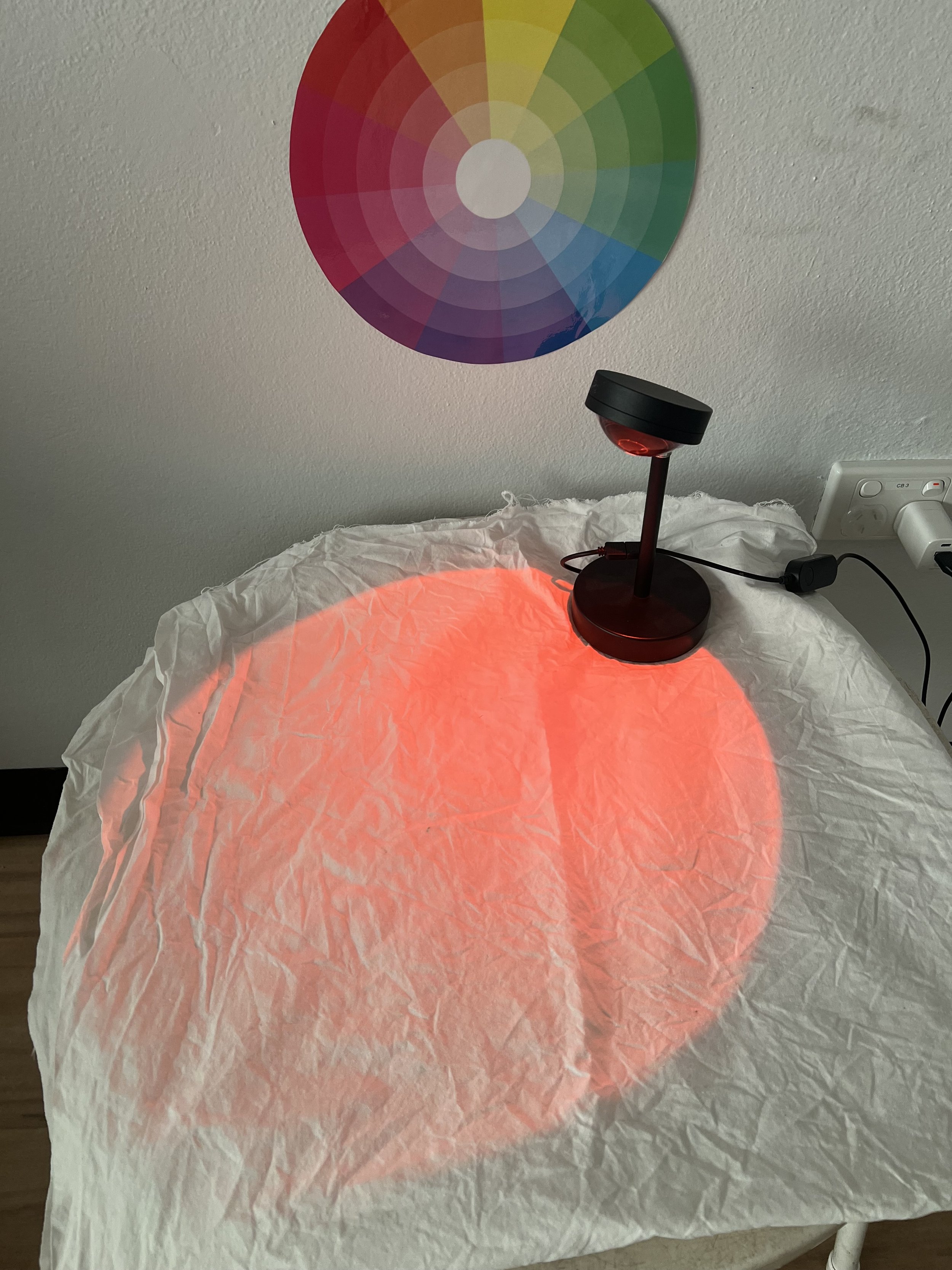 Color wheel on wall above a table with a red light source projecting a red circle onto a crinkled white paper surface.