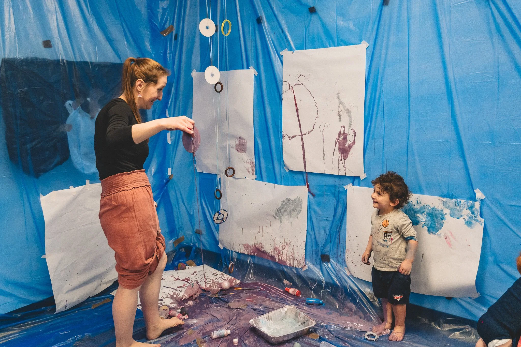 A woman and a young boy are arts and crafts in a room with blue plastic walls. The room is decorated with large paintings on paper, some covered in paint splatters and drips. The woman is holding a purple balloon, and the boy is smiling at her, with art supplies scattered on the floor.