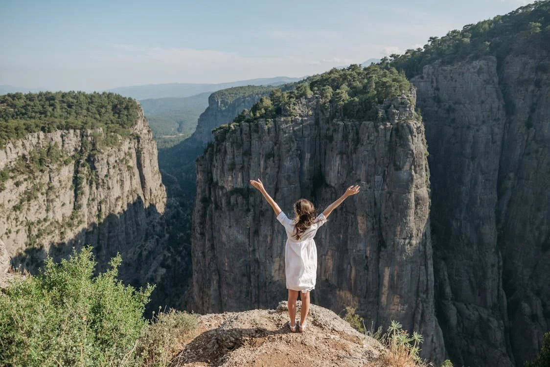 Woman in white dress standing in a rock formation shaped like a heart, overlooking a mountainous landscape under a clear blue sky.