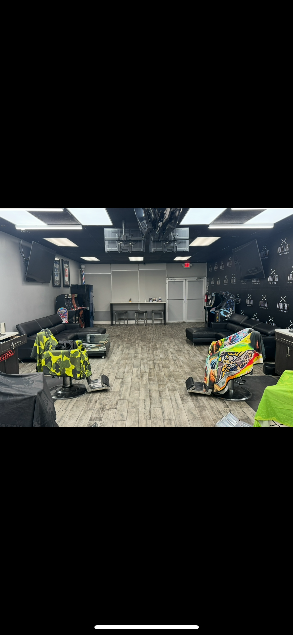 An empty indoor game lounge with black leather couches, arcade machines, and colorful chairs, featuring a gray wood floor, gray walls with framed art, and a black wall with the "WORLDKART" logo.
