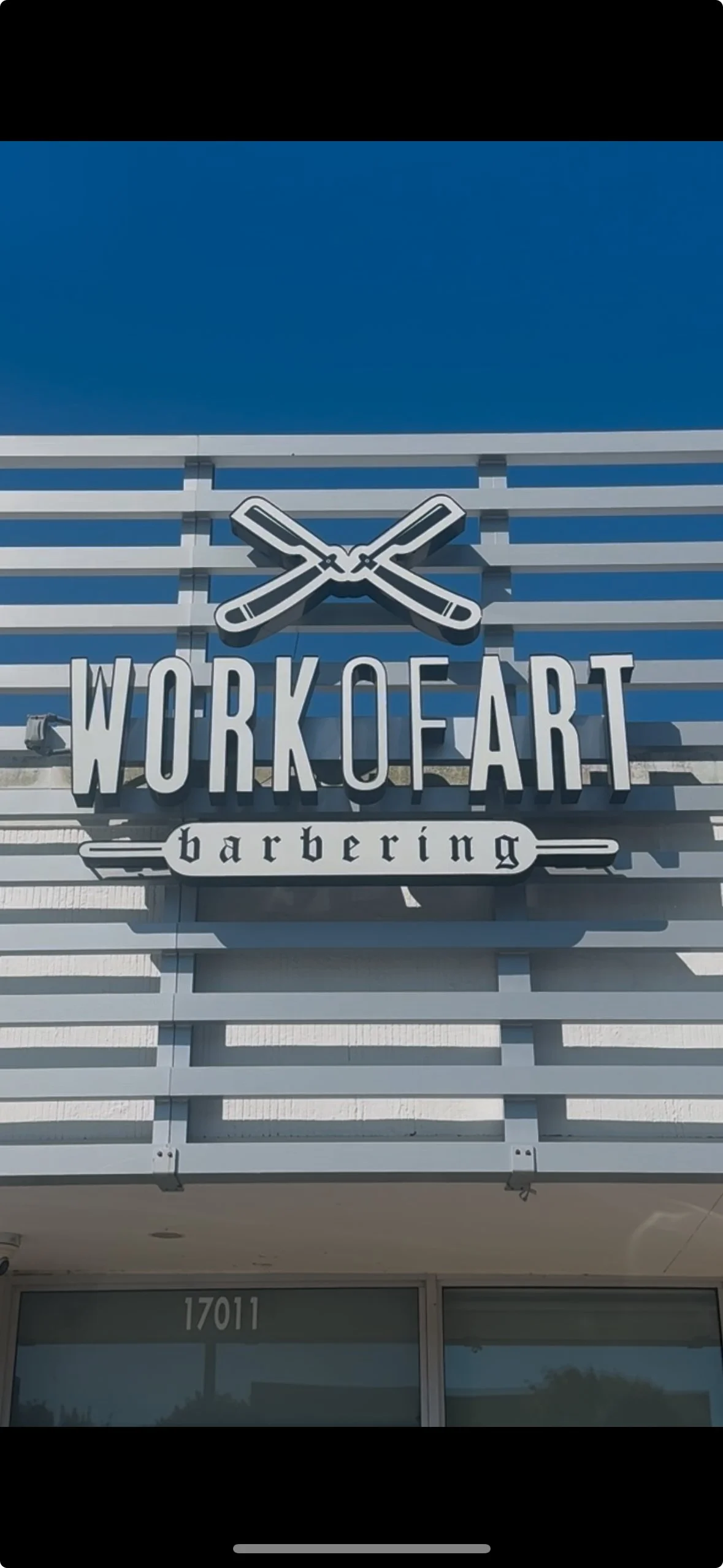 Sign for 'Work of Art' barbering with a stylized image of crossed straight razors above the text, on a modern building facade.