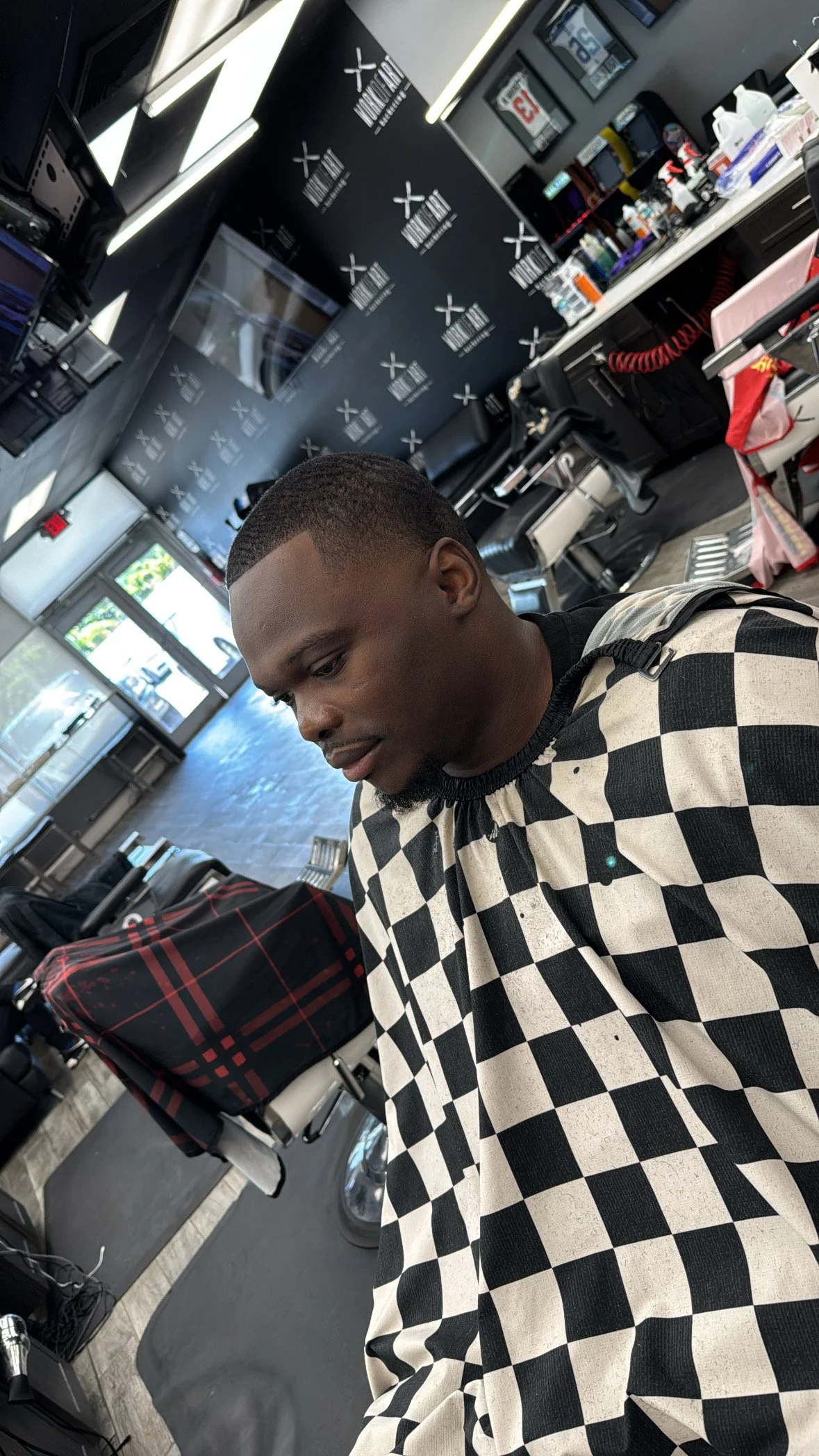 Young man sitting in a barbershop wearing a black and white checkered cape, with a haircut and reflective expression, with barber tools and chairs in the background.
