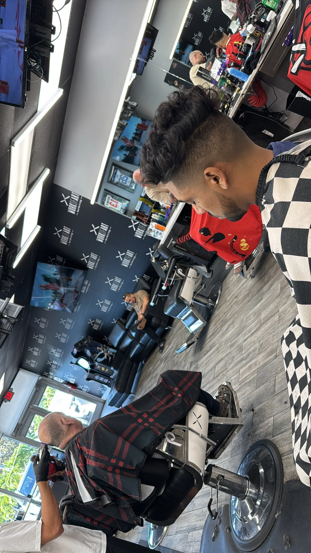 A man sitting in a barber chair getting a haircut, with a barber standing behind him, in a barbershop with black walls and a TV on the wall. The barbershop has a reception counter with various hair styling products and equipment, and there are other 