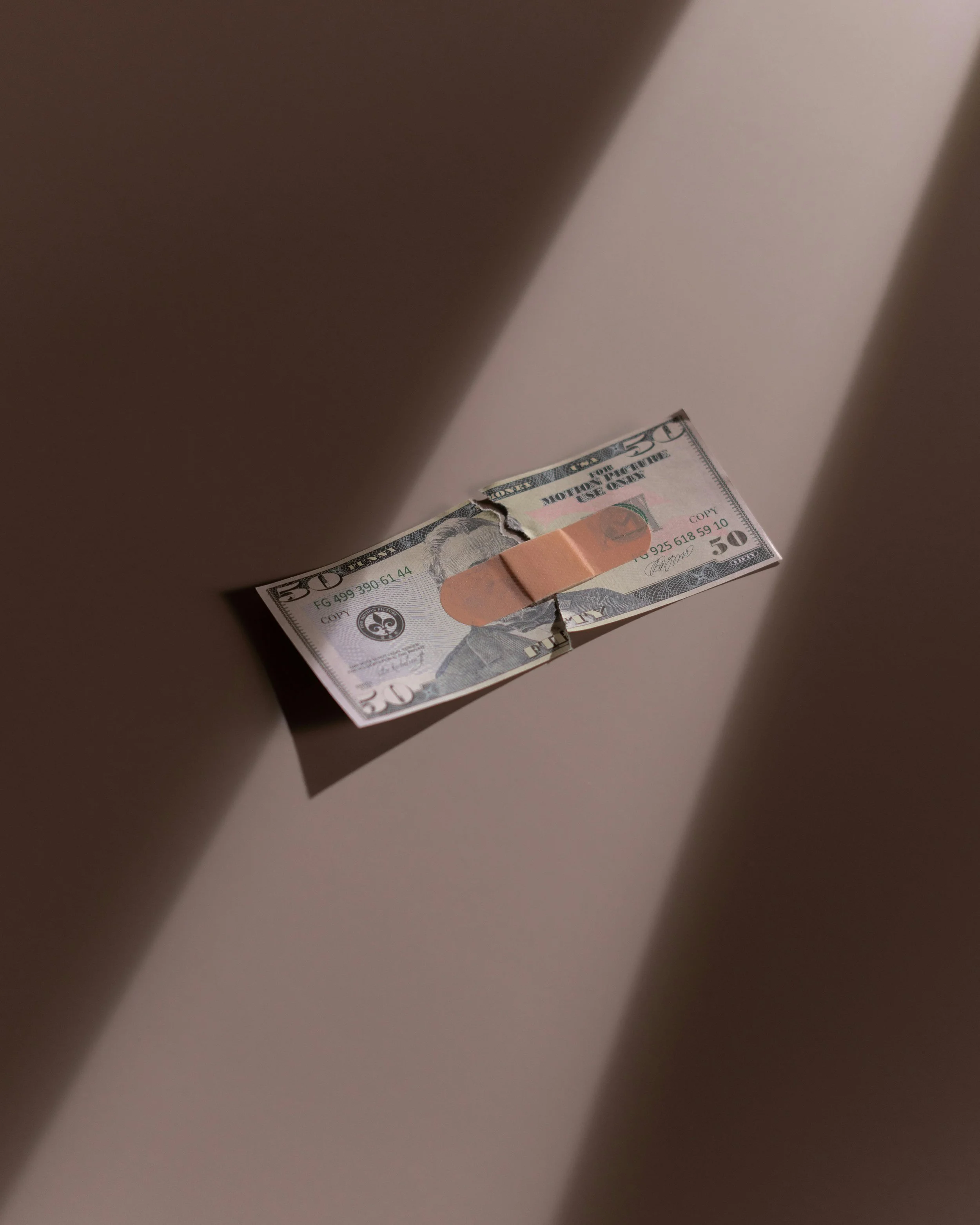 A fifty-dollar bill with a band-aid across the middle, casting a shadow on a plain background.