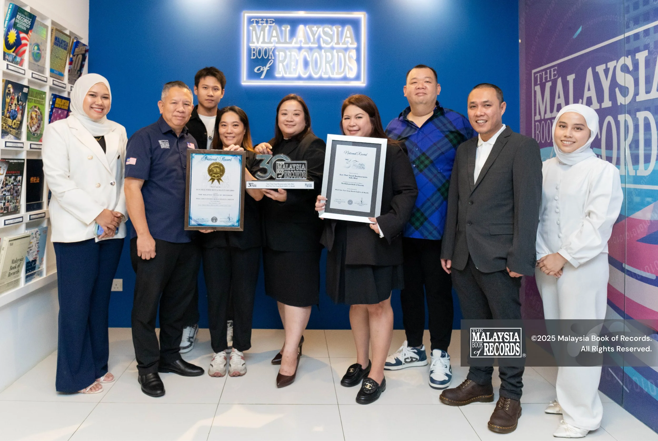 Ten people from Sun Thai Food family standing inside Malaysia Book of Records office, holding awards and certificates, with shelves of books on the left and a blue wall with illuminated sign reading 'The Malaysia Book of Records' behind them.