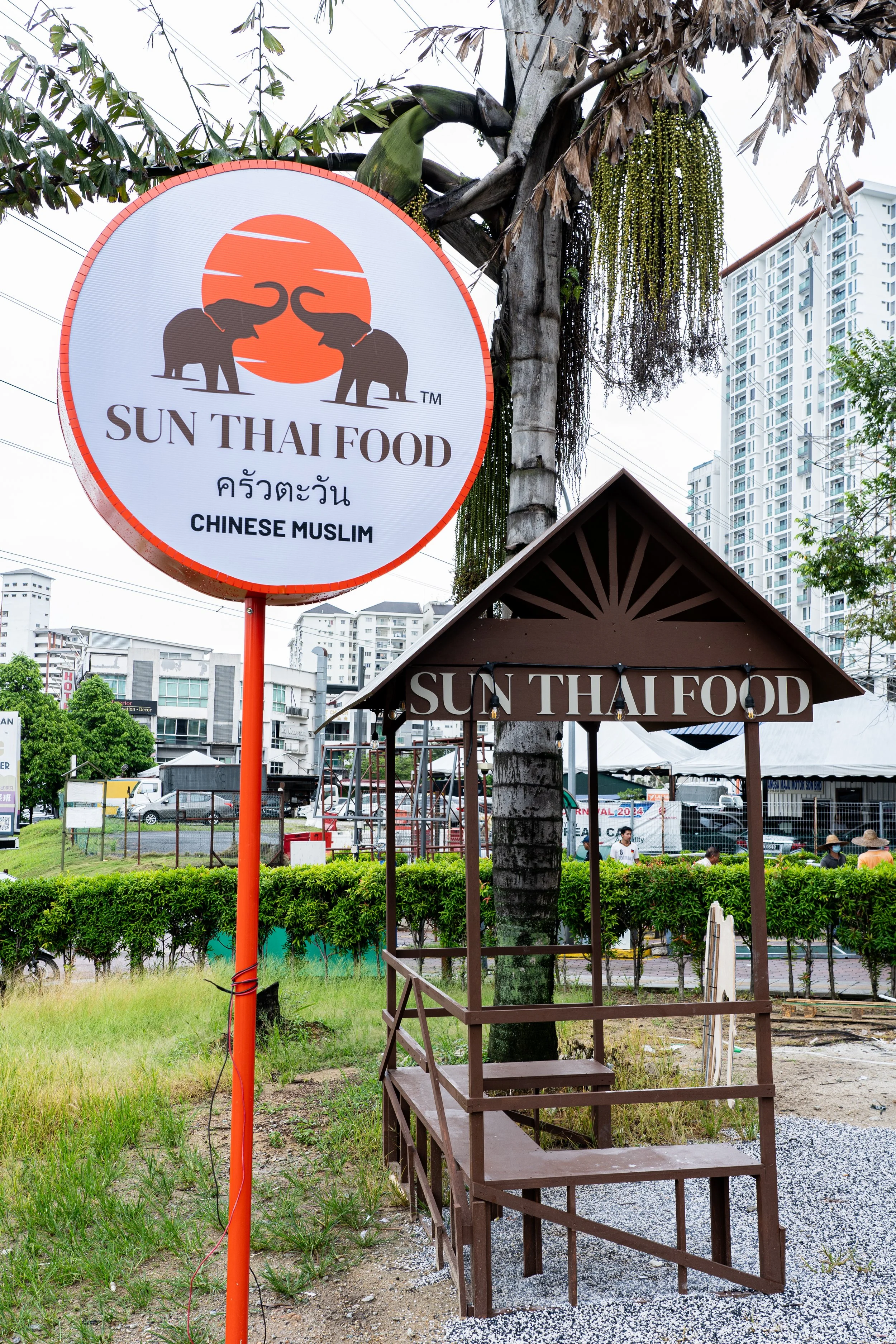 Sign for Sun Thai Food restaurant with an illustration of two elephants and a sunset, featuring Thai and English text, and a small wooden stand with a roof displaying the same restaurant name.