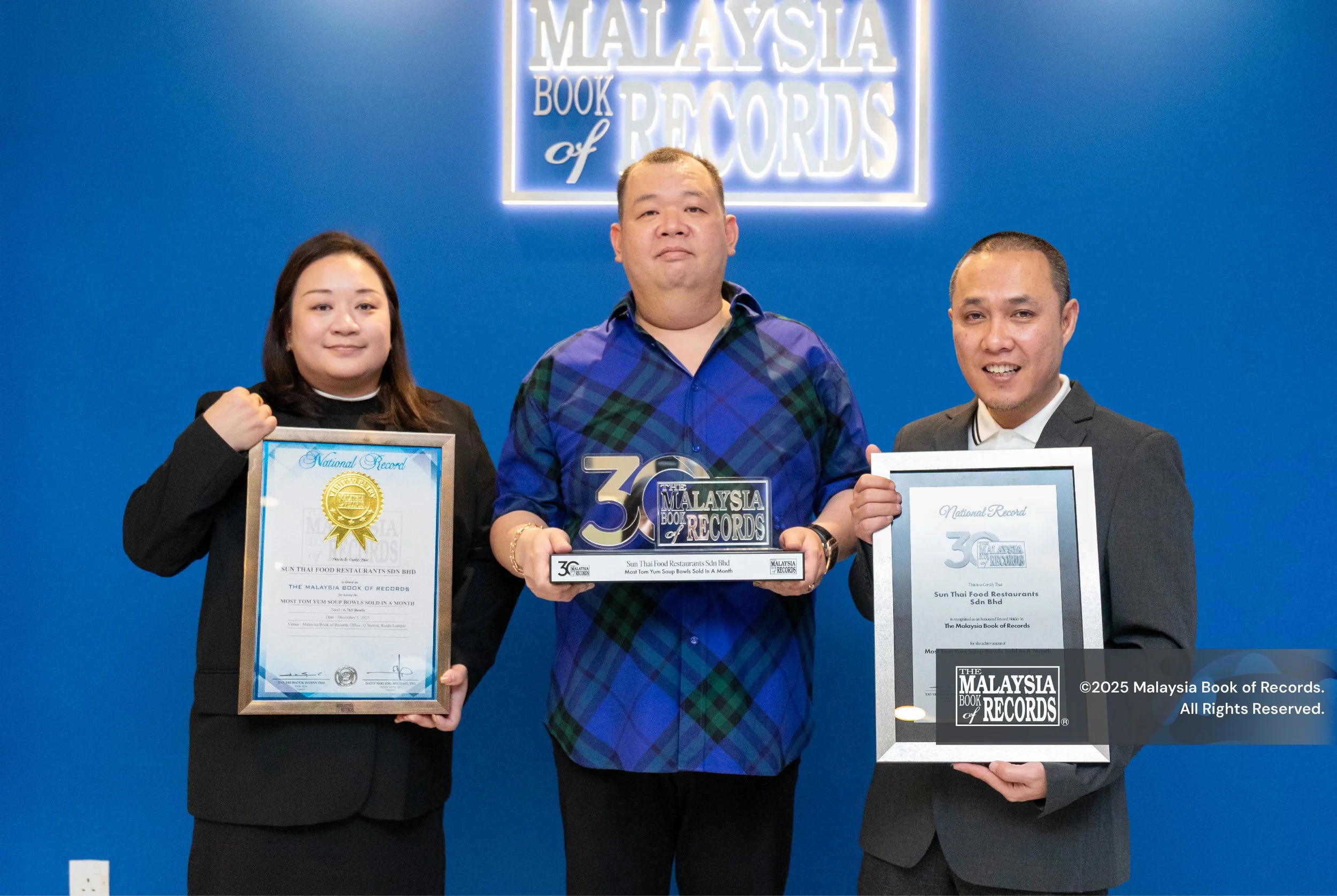 Three contributers of Sun Thai Food holding certificates and an award in front of a blue wall with a sign that reads 'Malaysia Book of Records.'