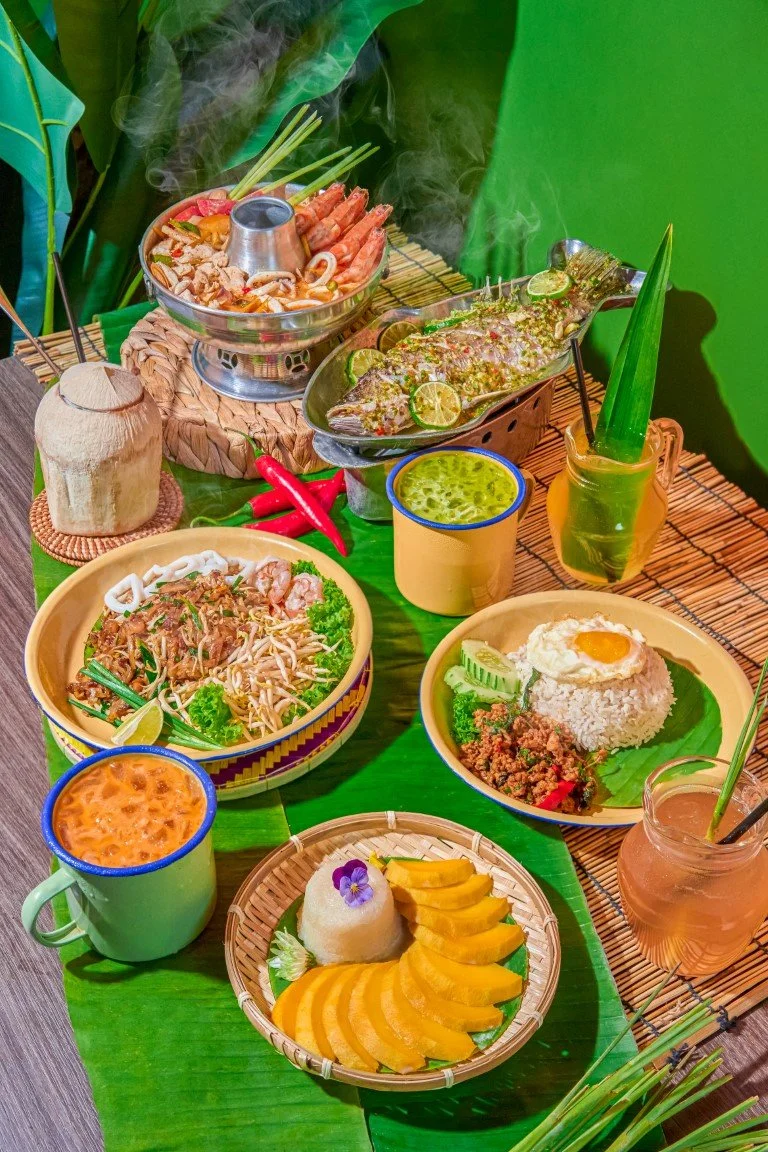 The picture of Sun Thai food restaurant. A vibrant spread of Thai food on a green table, including grilled fish with lemon, Tomyum seafood hotpot, Pad Thai noodles and various drinks and condiments.