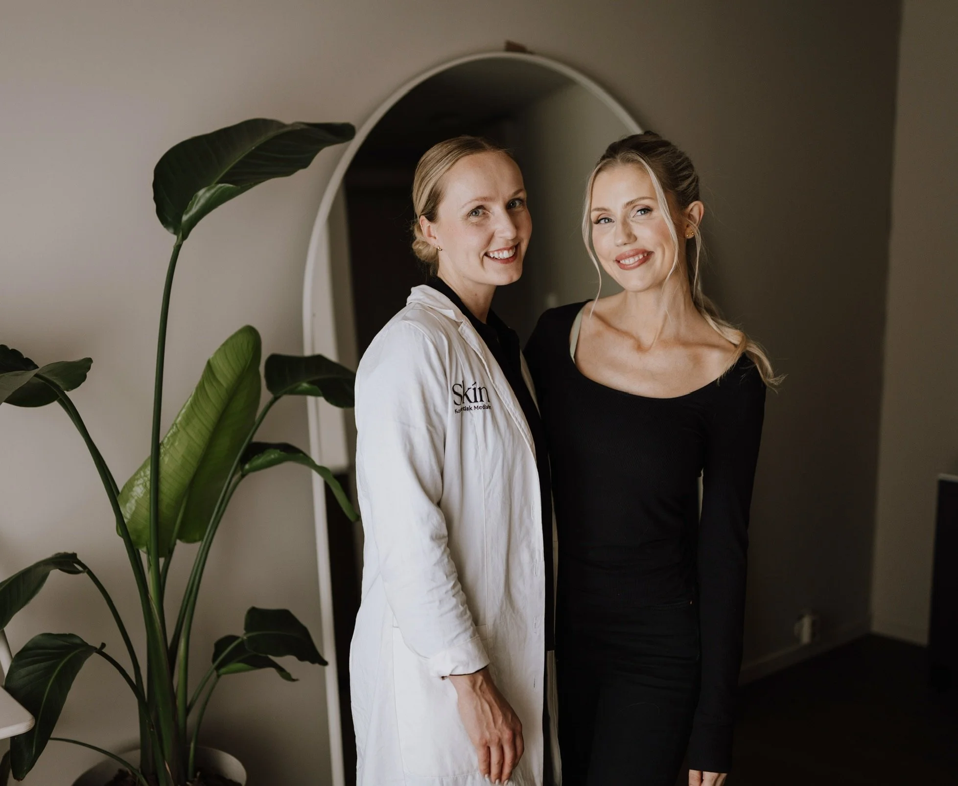 En estetisk lege og en komsetisk sykepleier smiler for kameraet, én i hvit labfrakk og én i svart kjole, står sammen innendørs foran et speil, med en stor grønn plante i forgrunnen.
