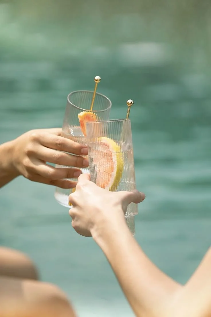 Due persone si abbracciano e brindano con bicchieri di vetro contenenti drinks con fette di agrumi, uno al limone e l'altro alla pompelmo, con sfondo di un corpo d'acqua.
