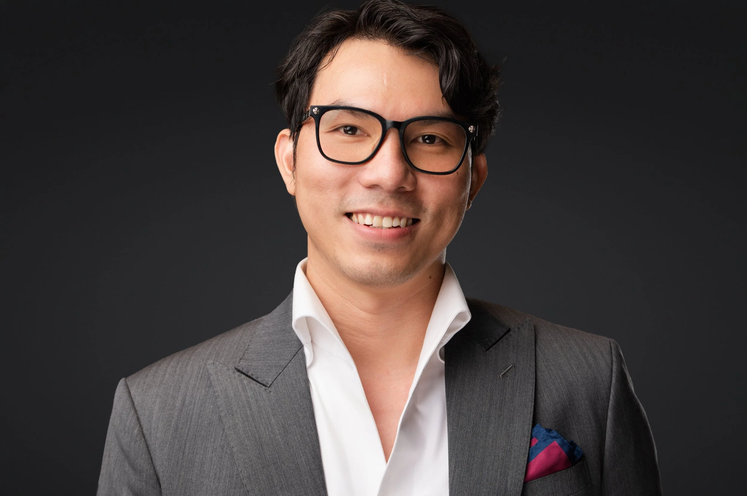Smiling man wearing glasses and a gray suit against a dark background