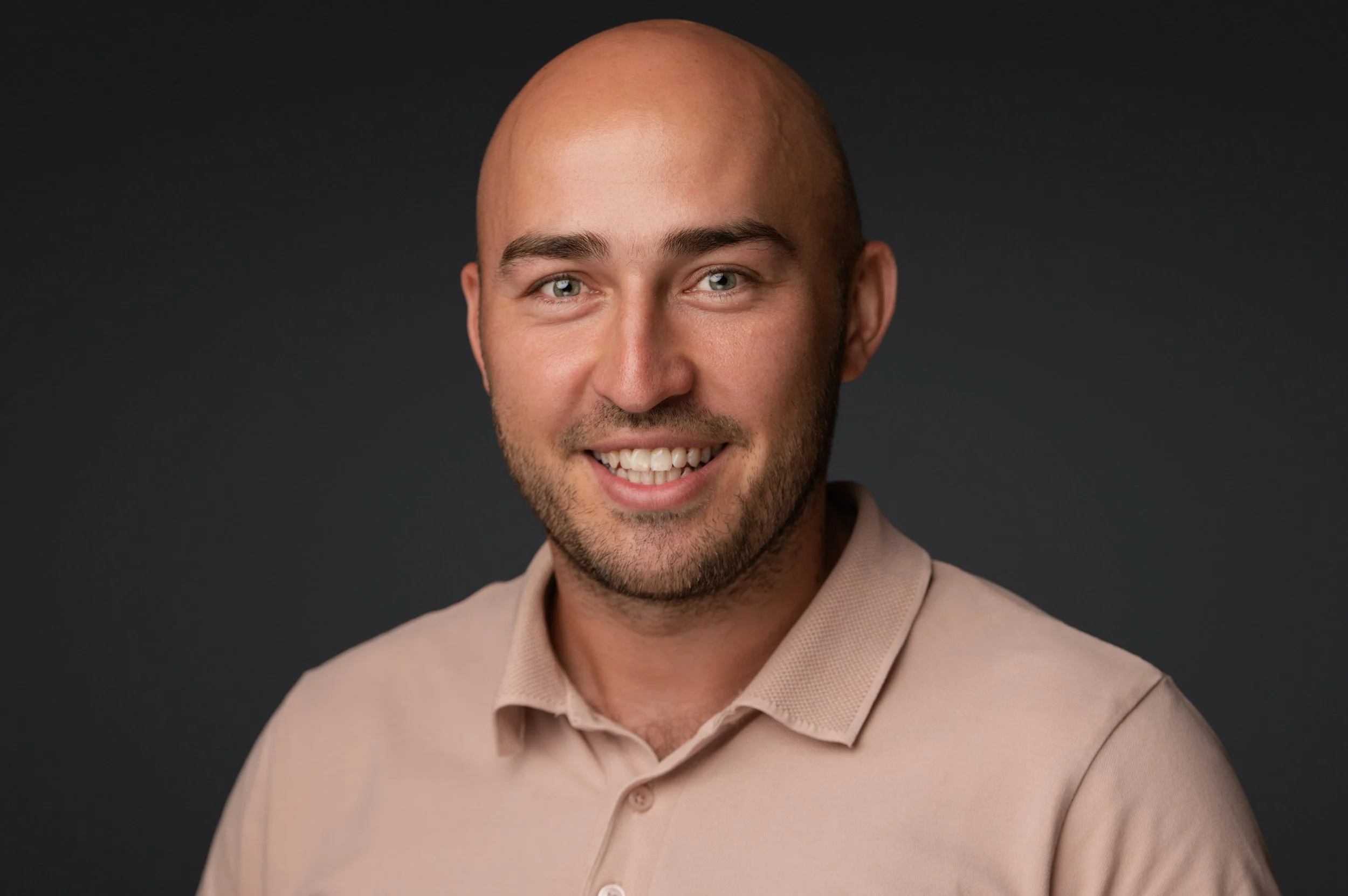 Smiling bald man in a beige polo shirt against a dark background.