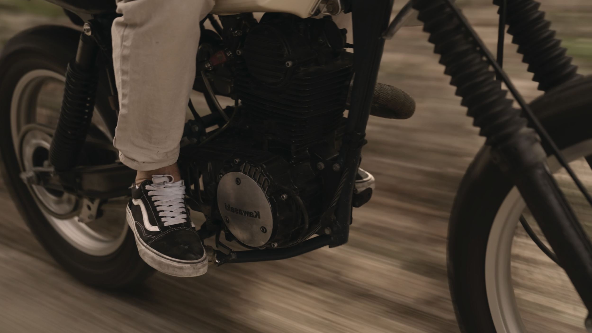 Close-up of a person riding a black Kawasaki vintage motorcycle, wearing black and white sneakers and beige pants, with a blurred background indicating motion.