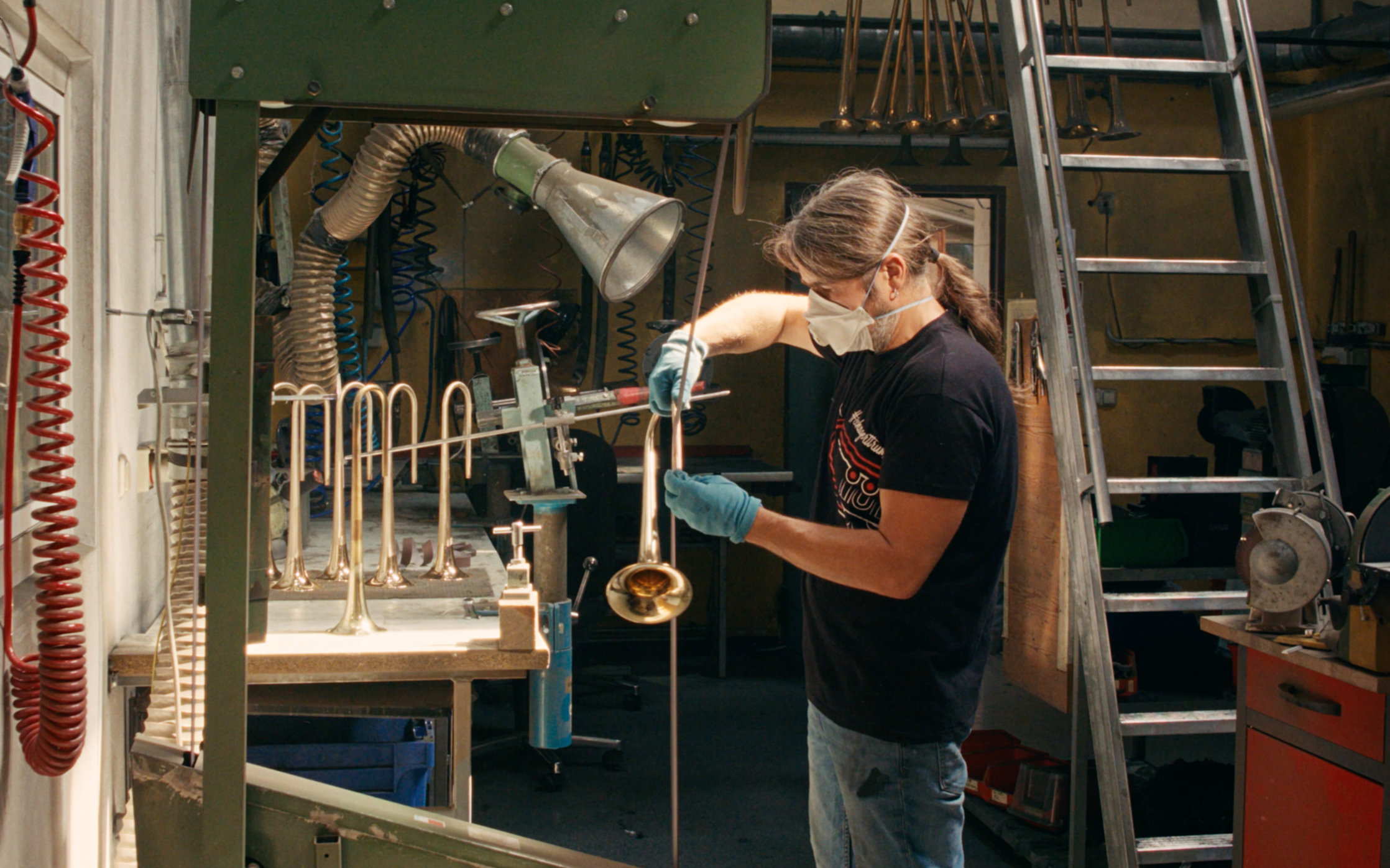 A Schagerl GmbH employee wearing a mask and gloves working on a brass instrument in a workshop with various tools and a ladder in the background.