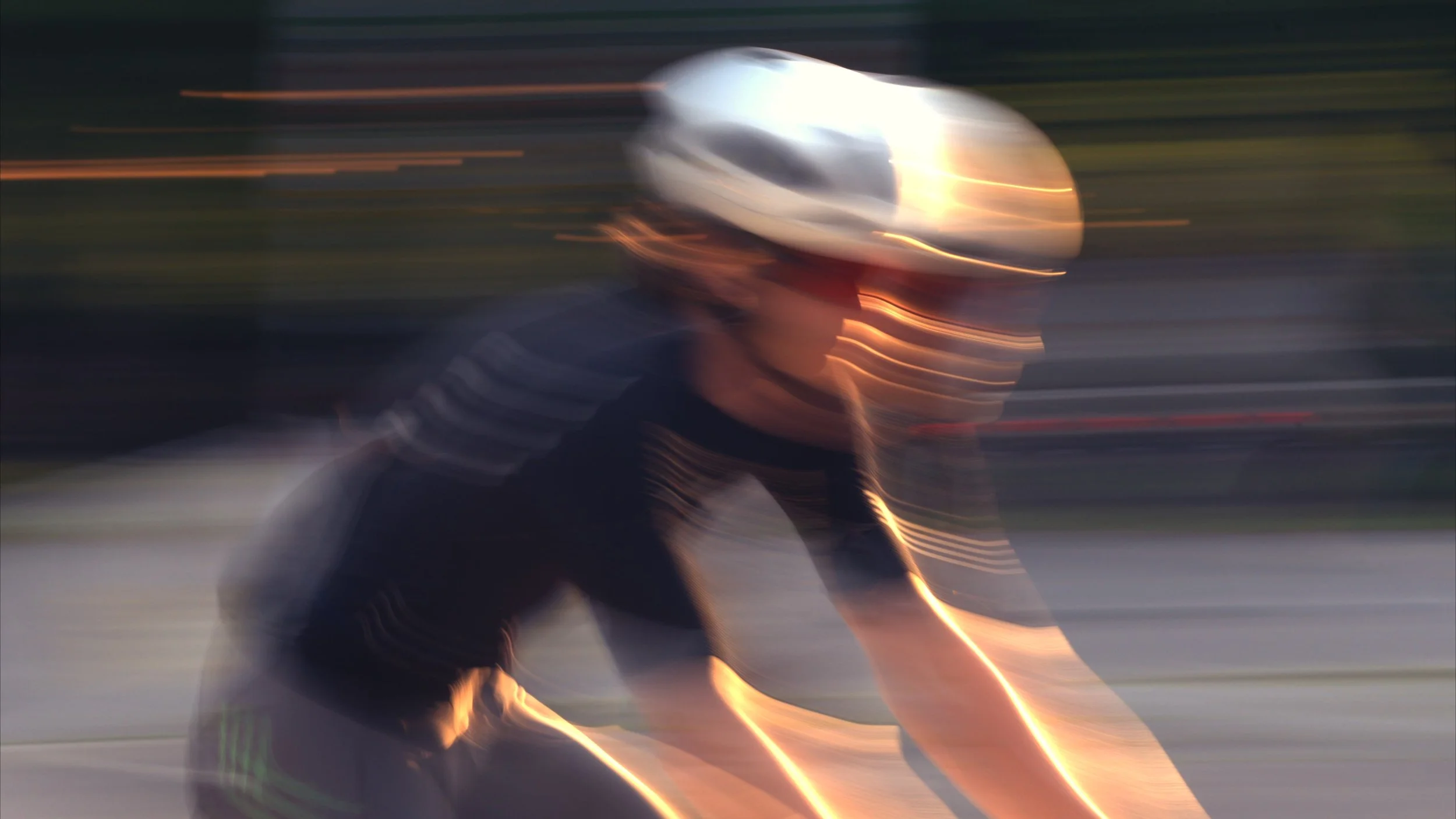 A person wearing a white helmet and a black shirt is riding a bicycle in motion, with a blurred background indicating fast movement at sunset or dusk.