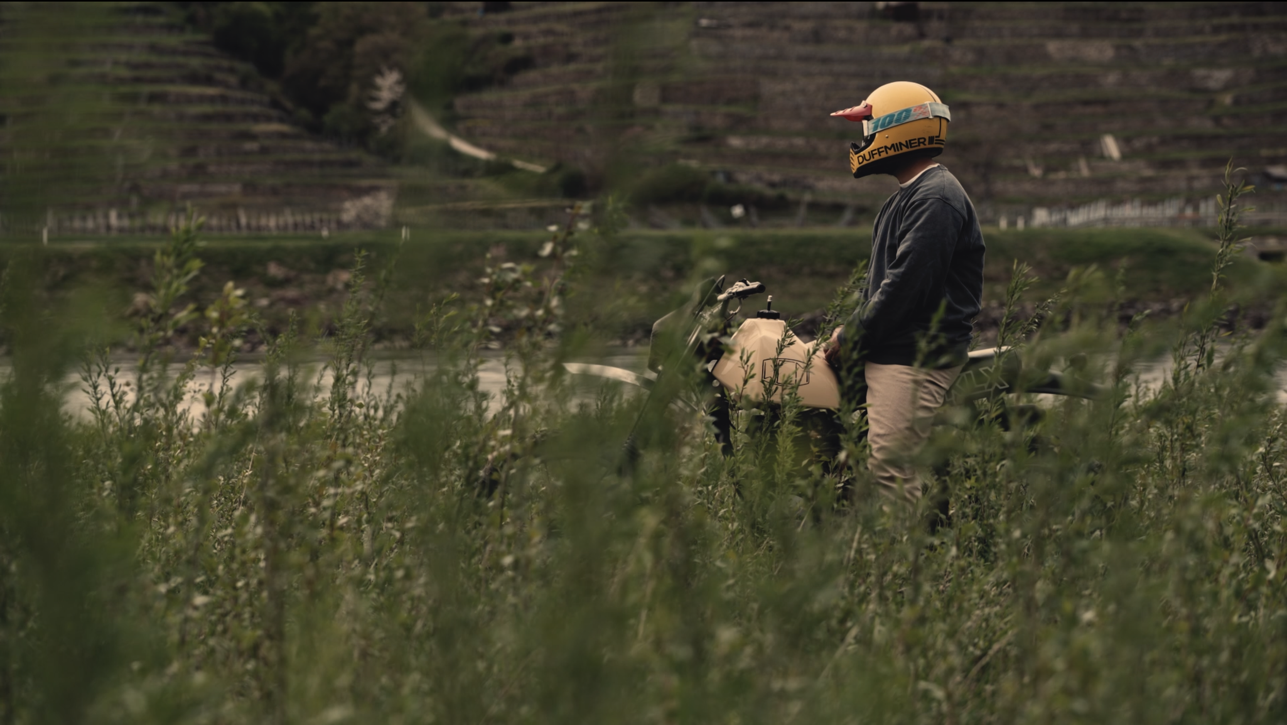 A person wearing a helmet and dark jacket standing next to a motorcycle in a green, hilly landscape with terraced fields in the background.