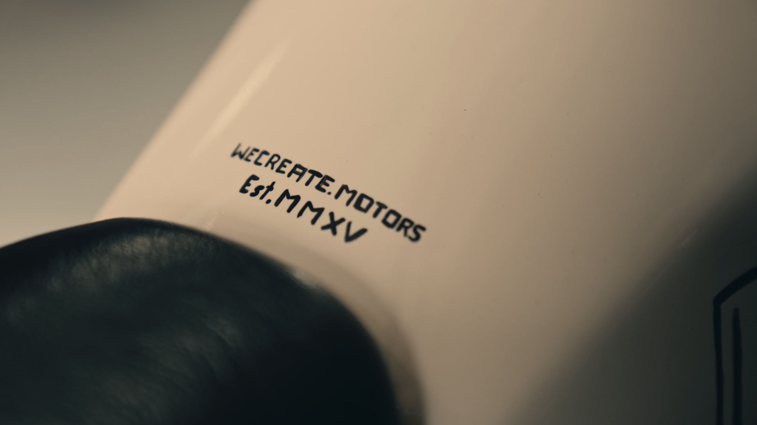Close-up of the text 'WE CREATE MOTORS Est. M MXV' on a beige machine part, with a black rounded object in the foreground.