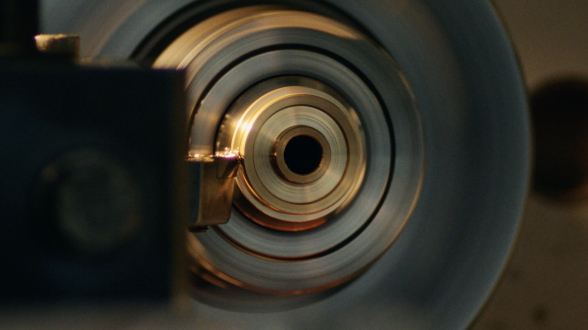 Close-up view of a metallic machine part with concentric circles, a small spark or reflection near the edge, and blurred surrounding components.
