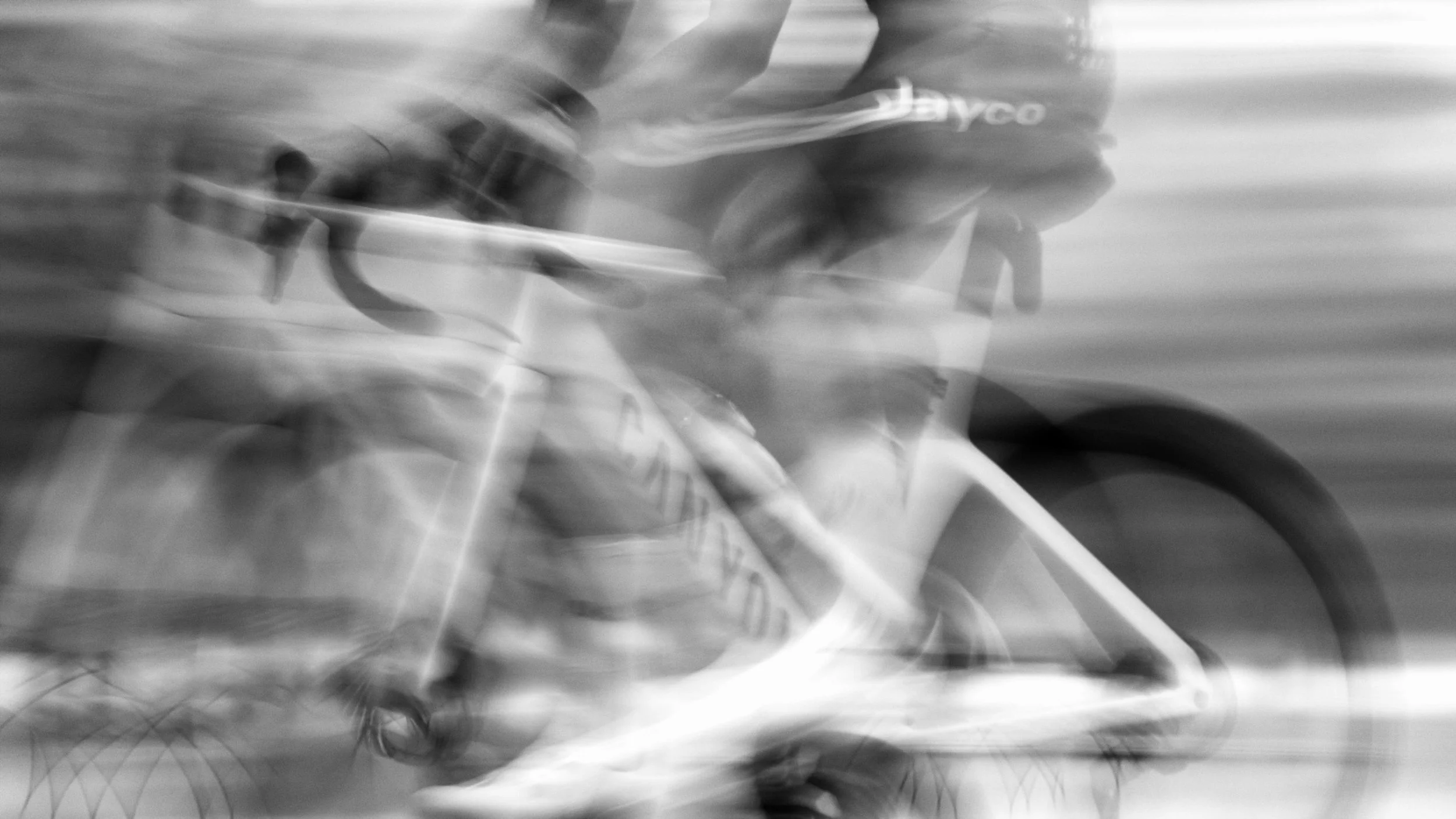 Blurred black and white photo of a person riding a bicycle, motion obscures details, with part of the bicycle and rider's face partially visible.