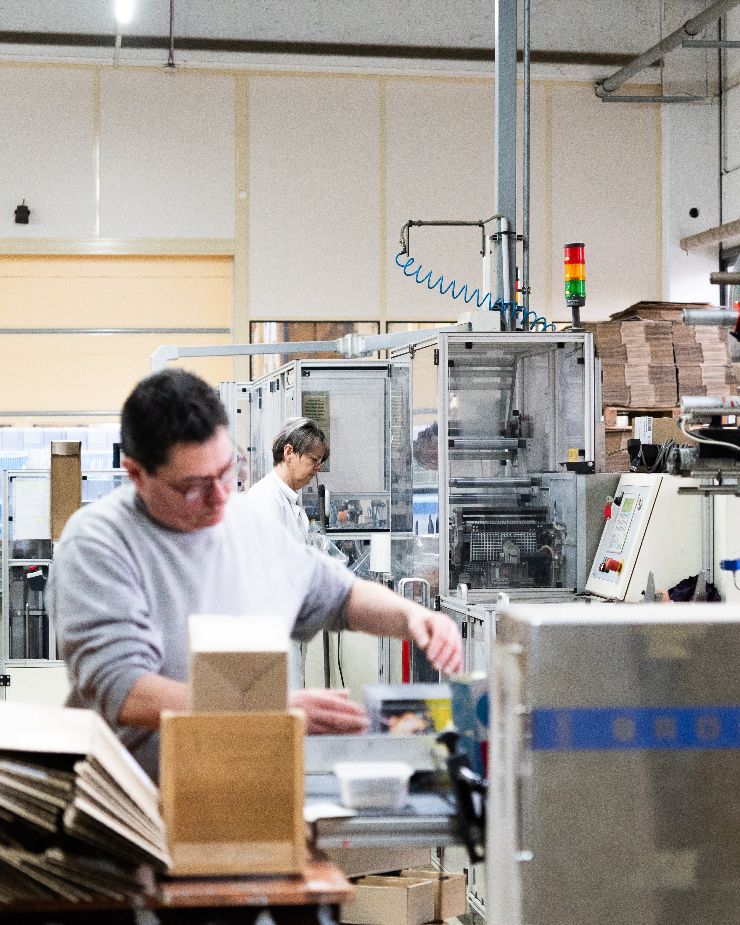 Deux personnes travaillent dans une usine ou une ligne de production, entourées de machines et de piles de carton.
