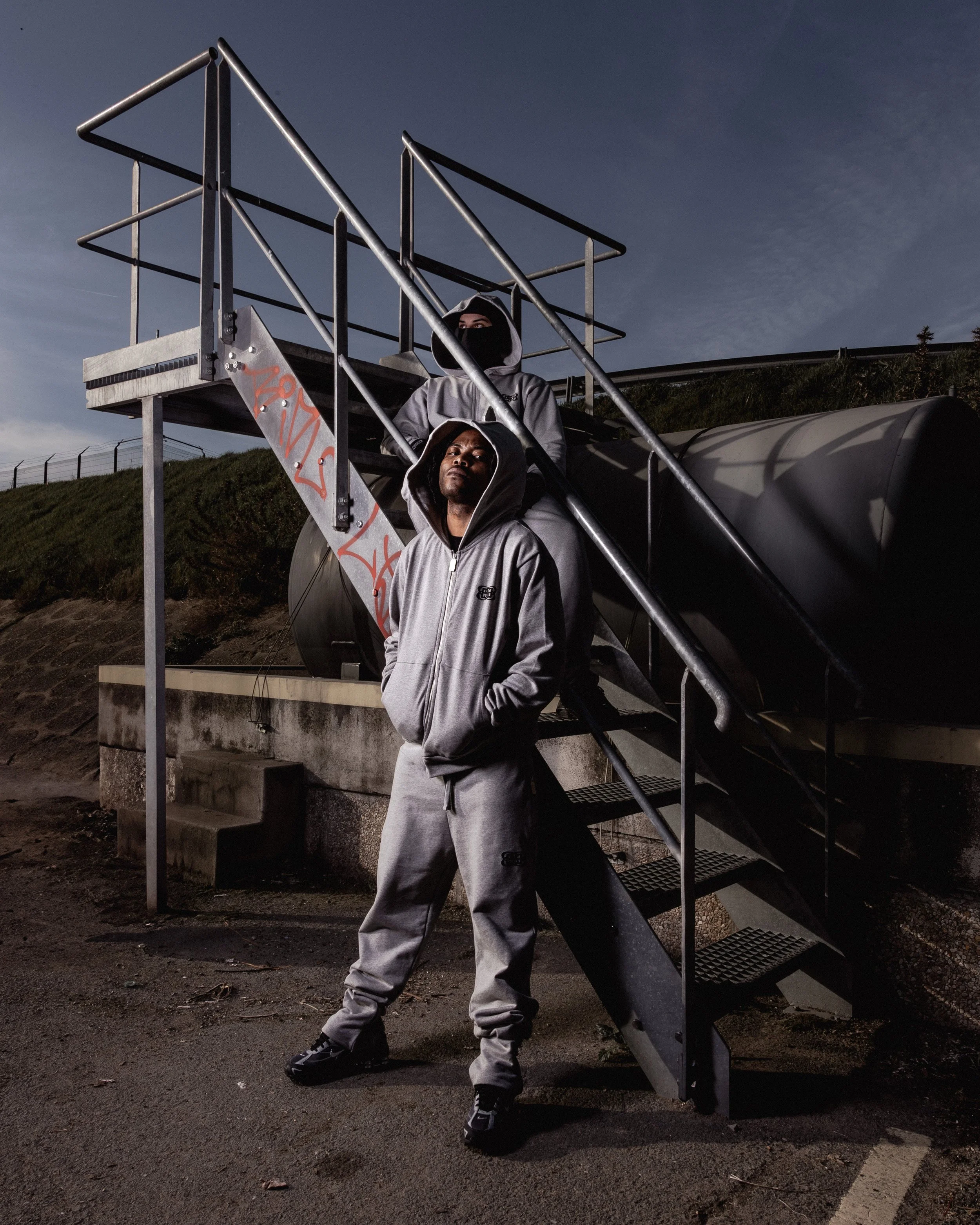 Deux personnes en vêtements de sport gris, avec capuches, posent devant une structure en métal et un réservoir d'eau, en extérieur au crépuscule ou à l'aube.