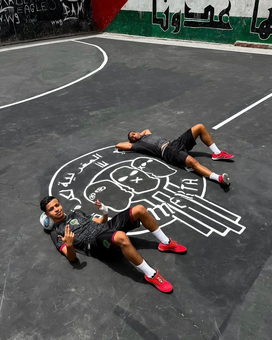 Deux jeunes hommes en tenue de sport reposent sur un sol peint d'un terrain de sport, avec un logo représentant un crâne stylisé en noir et blanc. Ils portent des shorts noirs avec des logos verts, des T-shirts noirs, des chaussettes blanches, et des chaussures rouges. Un d'eux sourit et fait un geste avec la main, tandis que l'autre est allongé avec les bras derrière la tête. Le terrain est entouré de murs peints avec des graffitis et des inscriptions en arabe.