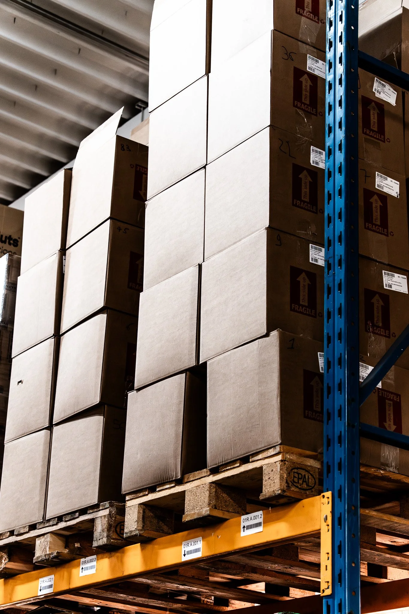 Boîtes en carton empilées sur une palette dans un entrepôt, avec des étiquettes et à côté d'une étagère en métal bleu.