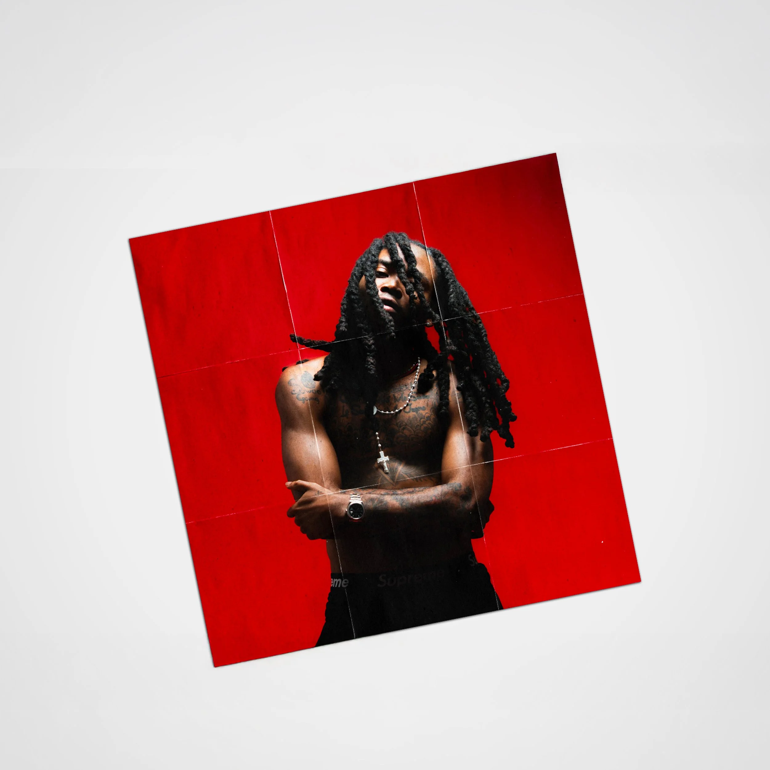 Un homme nu avec des tatouages, portant une chaîne avec une croix, se tient contre un fond rouge.