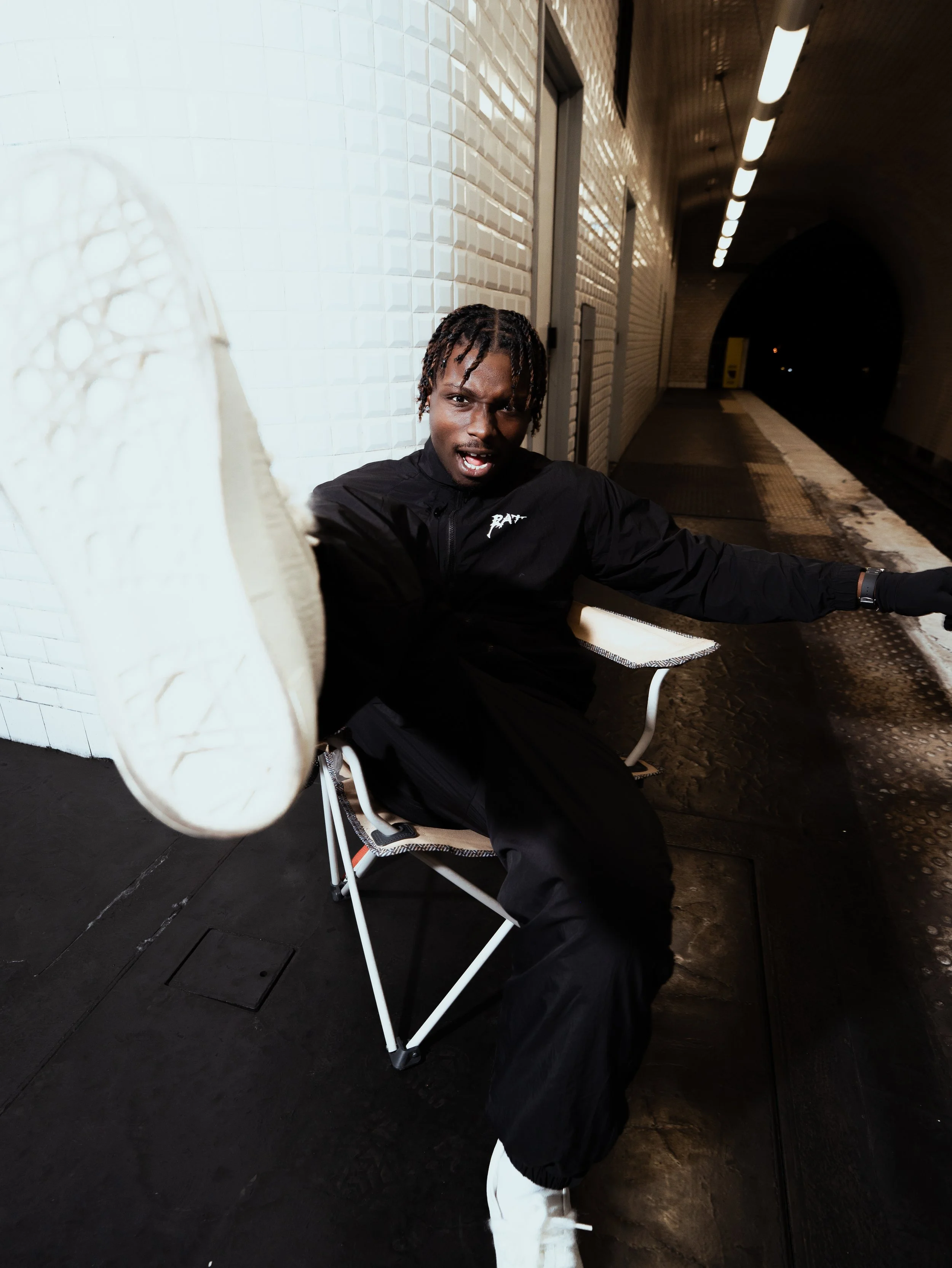 Un homme en vêtements noirs assis sur une chaise dans un tunnel, faisant un geste vers l'appareil photo, avec une expression intense.