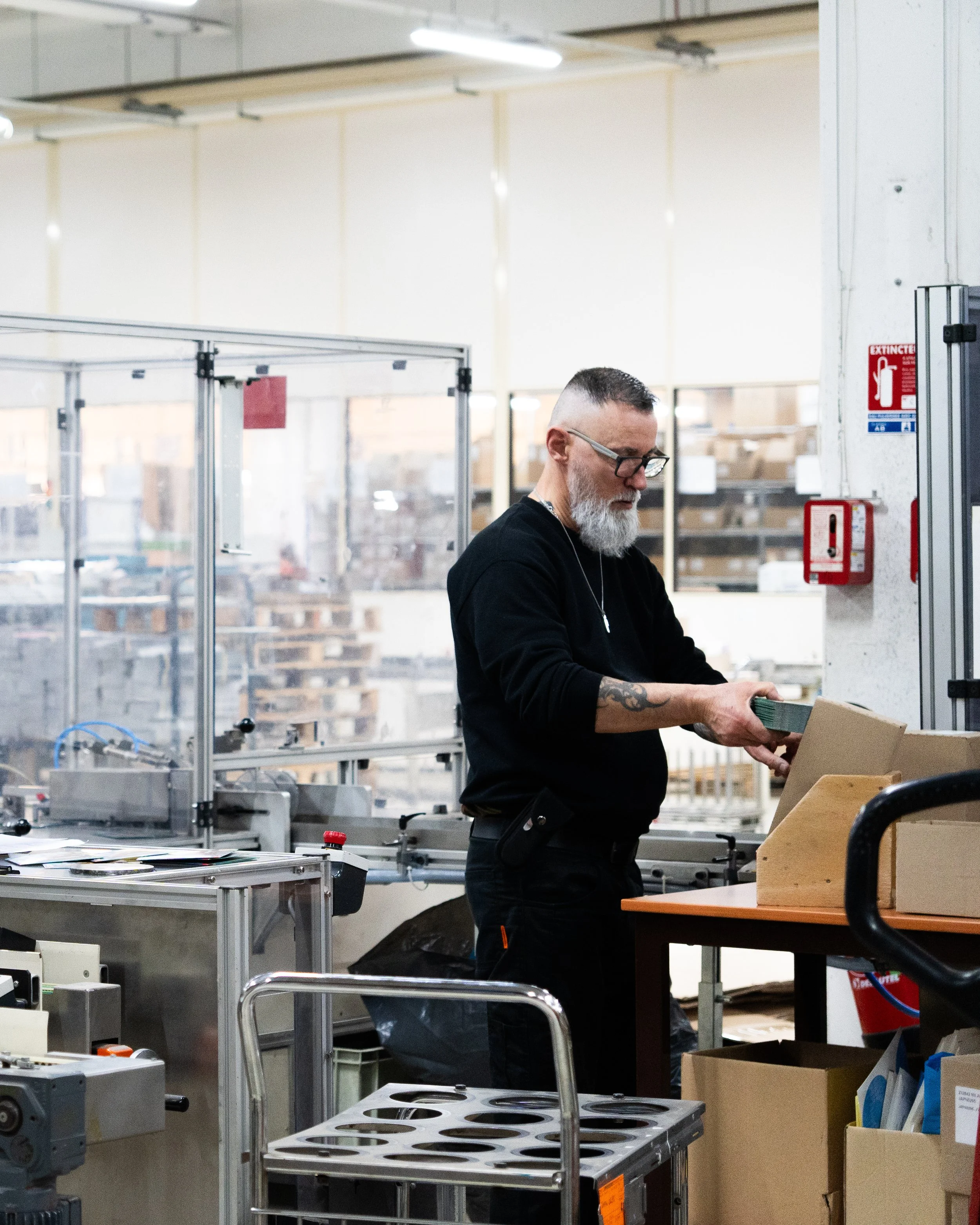 Un homme avec une barbe grise, lunettes, portant un vêtement noir, travaille dans un atelier industriel, manipulant des cartons ou des pièces de matériel.